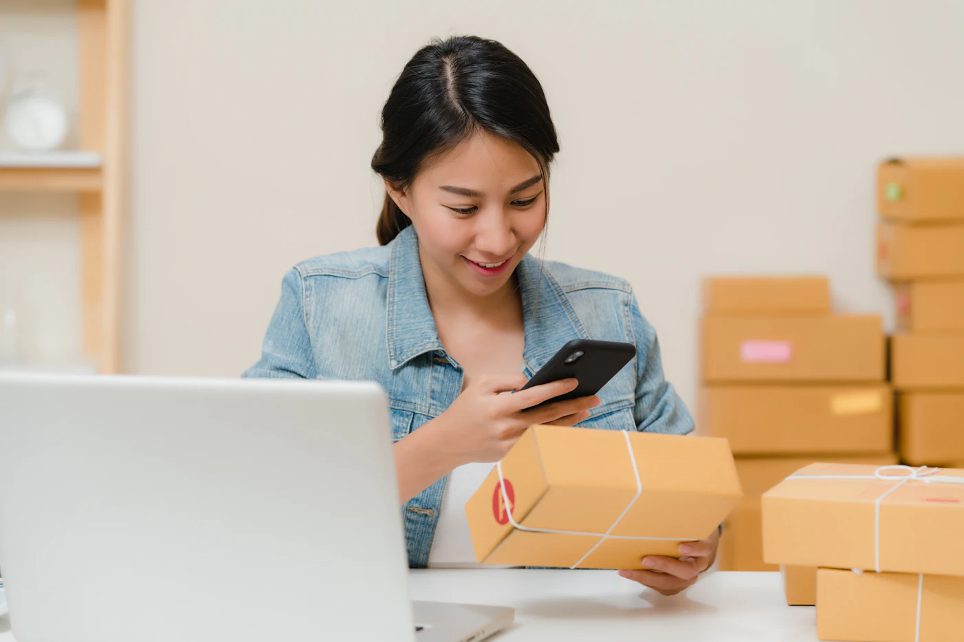 Agence Google Shopping : une commerçante en ligne vérifie un colis avec son téléphone, près de son ordinateur et de cartons d’expédition